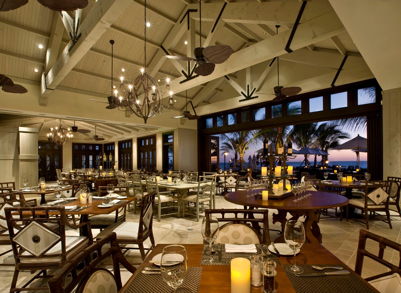 Beachfront resort restaurant at dusk with white timber-frame ceiling, candlelit tables, and ocean view