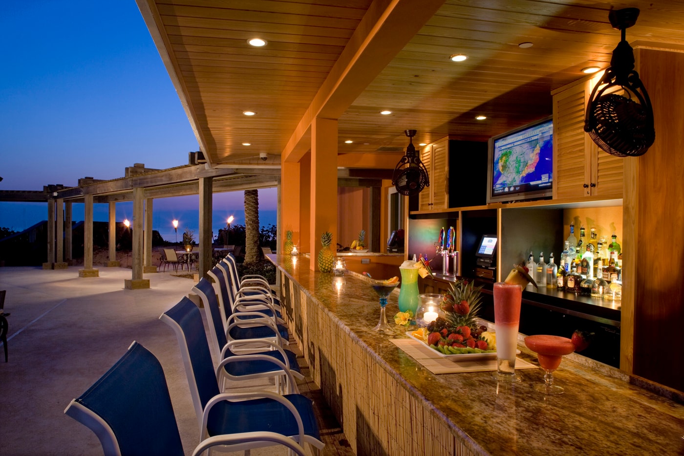 Outdoor beachfront tiki bar at dusk with wood ceiling, blue bar stools, tiki torches, and ocean view