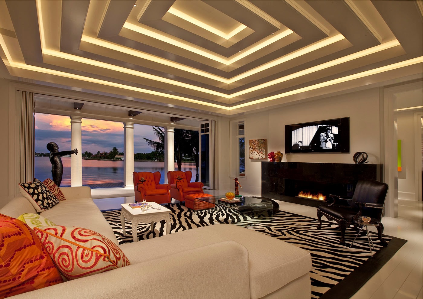 Dramatic waterfront living room at dusk with coffered LED ceiling, zebra rug and orange chairs