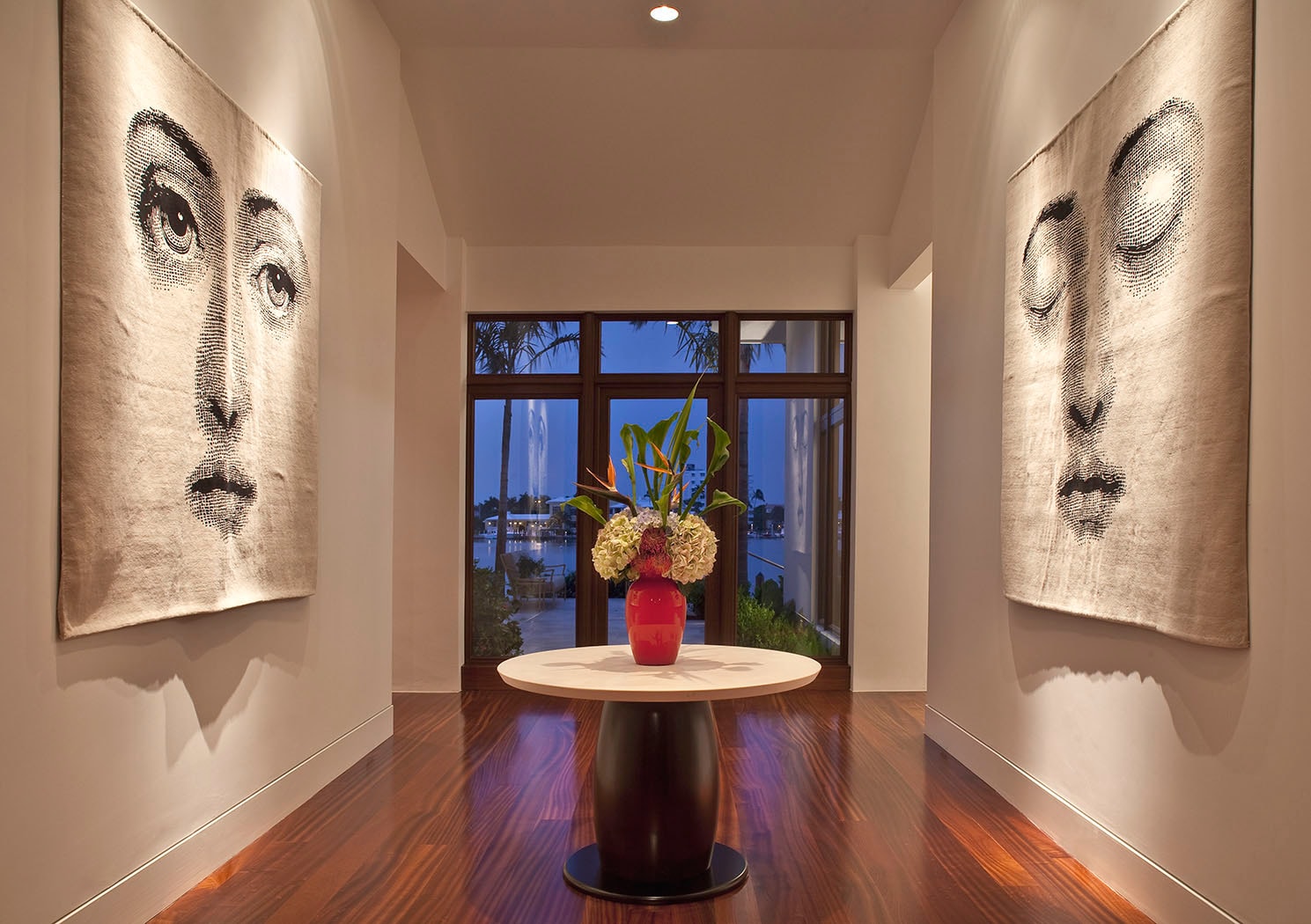 Minimalist hallway with two large Fornasetti face tapestries flanking a round pedestal table with flowers